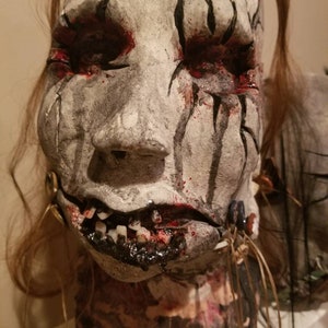 May include: A white and gray mask with black lines and red paint. The mask has a cracked and weathered appearance. The mask has a mouth with teeth and a black substance dripping from it. The mask is on a mannequin head with brown hair.