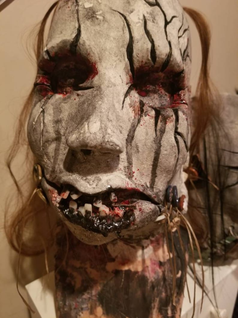 May include: A white and gray mask with black lines and red paint. The mask has a cracked and weathered appearance. The mask has a mouth with teeth and a black substance dripping from it. The mask is on a mannequin head with brown hair.