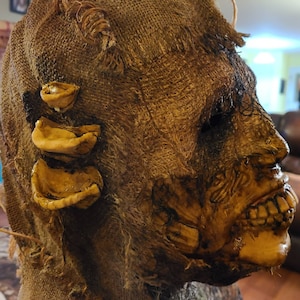 May include: A burlap-wrapped Halloween mask with a grotesque, decaying face and exposed teeth. The mask has three yellow, fleshy appendages on the side.