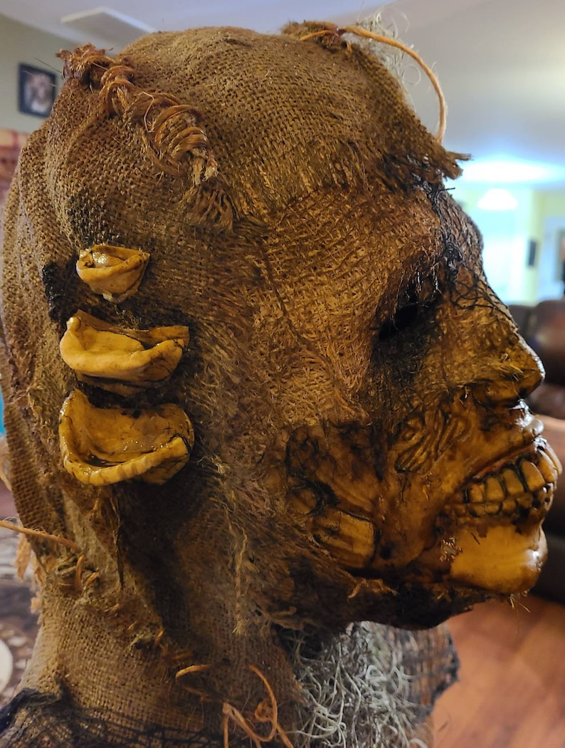May include: A burlap-wrapped Halloween mask with a grotesque, decaying face and exposed teeth. The mask has three yellow, fleshy appendages on the side.