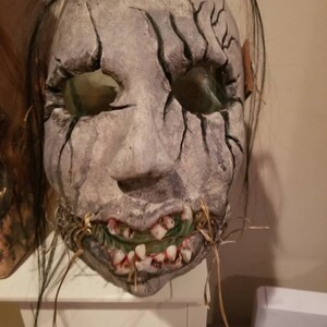 May include: A creepy, realistic-looking mask with a cracked, gray face, black hair, and a stitched-up mouth with visible teeth. The mask is made of papier-m&acirc;ch&eacute; and has a distressed, weathered look.