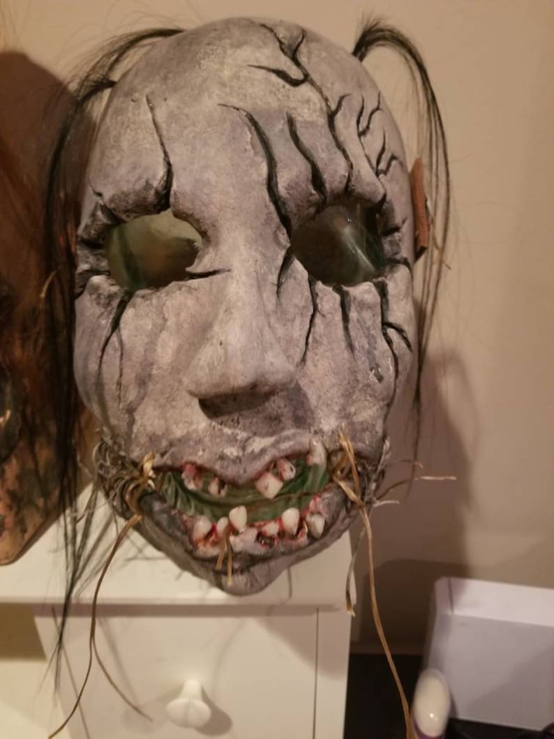 May include: A creepy, realistic-looking mask with a cracked, gray face, black hair, and a stitched-up mouth with visible teeth. The mask is made of papier-m&acirc;ch&eacute; and has a distressed, weathered look.
