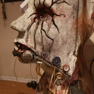 May include: A white, plaster-like mask with a red eye and black veins. The mask has a stitched mouth with black and brown buttons attached to the chin.