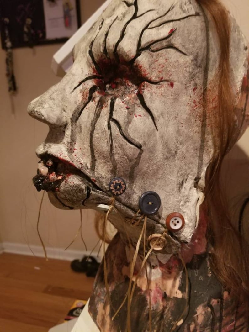 May include: A white, plaster-like mask with a red eye and black veins. The mask has a stitched mouth with black and brown buttons attached to the chin.