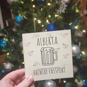 Op de afbeelding: Een crèmekleurige kaart met de tekst "ALBERTA BREWERY PASSPORT" en een illustratie van een bierpul, gehouden voor een kerstboom versierd met blauwe en zilveren ballen en lichtjes.