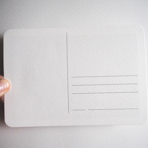 May include: A blank, off-white postcard with rounded corners. The back of the card features vertical and horizontal lines for writing addresses and a message. The card is held by a person's hand.