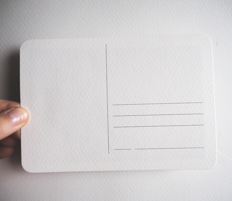 May include: A blank, off-white postcard with rounded corners. The back of the card features vertical and horizontal lines for writing addresses and a message. The card is held by a person's hand.