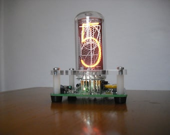 May include: A Nixie tube clock displaying the number 5 in a warm orange glow. The tube is encased in clear glass and mounted on a green circuit board with a clear acrylic base. The clock sits on a brown wooden surface.