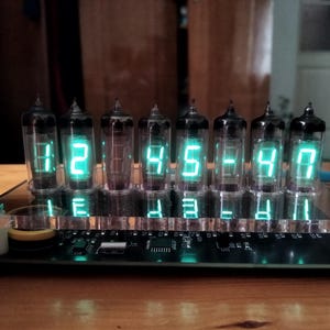 May include: A digital clock with seven glass tubes displaying the time 12:45:47 in bright green numerals. The clock sits on a black base with a clear acrylic front panel, and is on a wooden surface.