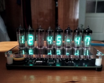 May include: A digital clock with seven glass tubes displaying the time 12:45:47 in bright green numerals. The clock sits on a black base with a clear acrylic front panel, and is on a wooden surface.
