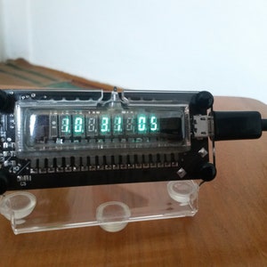 May include: A black electronic device with a clear plastic cover displays the numbers "30.383.05" in green. The device is connected to a black cable and rests on a clear plastic stand. The device has black knobs on each corner.