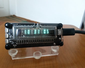 May include: A black electronic device with a clear plastic cover displays the numbers "30.383.05" in green. The device is connected to a black cable and rests on a clear plastic stand. The device has black knobs on each corner.