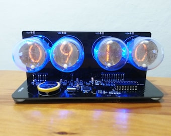 May include: A black digital clock with four glowing Nixie tubes displaying the time as 09:10. The clock has a blue LED backlight and sits on a wooden surface. The clock's design is retro and futuristic.