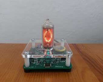 May include: A Nixie tube clock with a clear acrylic top and base. The orange-glowing tube displays a number. The clock is mounted on a green circuit board with the text "TRCorp".