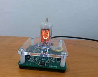 May include: A small electronic device featuring a glowing orange Nixie tube display. The tube is encased in clear glass and mounted on a green circuit board with a clear acrylic cover. The device is on a wooden surface.