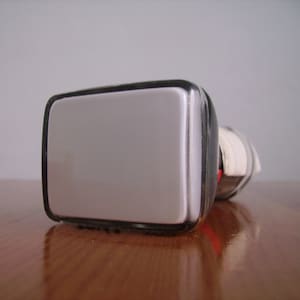 Puede incluir: Un objeto rectangular plateado con un borde negro. El objeto tiene una parte superior redondeada y está colocado sobre una superficie de madera. El fondo es de color gris claro liso. El objeto parece ser un espejo.