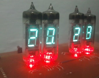 May include: A close-up of a digital clock displaying the time 2:18. The clock uses four Nixie tubes with a retro aesthetic. The digits glow bright teal, and the base emits a red light. The clock is mounted on a brown circuit board.