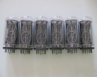 May include: Six vintage vacuum tubes with clear glass enclosures and intricate internal wire structures. Each tube has a series of pins at the base and a rounded top. The tubes are arranged in a row against a white background.