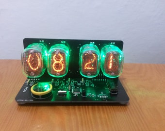 May include: A digital clock with a black base and four glowing orange digits encased in glass tubes. The tubes are illuminated with a green glow. The clock displays the time "08:21". The clock sits on a wooden surface.
