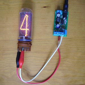 Pode incluir: Um tubo Nixie vintage exibindo o número 4 em um brilho laranja quente. O tubo está conectado a uma pequena placa de circuito com fios e clipes de jacaré. Os componentes estão sobre uma superfície de madeira.