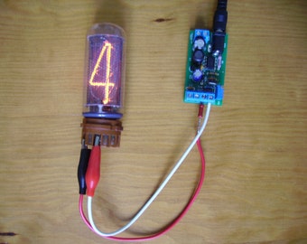 May include: A vintage Nixie tube displaying the number 4 in a warm orange glow. The tube is connected to a small circuit board with wires and alligator clips. The components are set against a wooden surface.