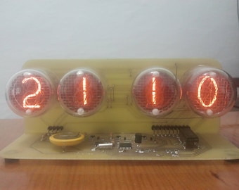 May include: A digital clock displaying the time 20:00. The clock features four glowing orange-lit vacuum tubes, mounted on a yellow circuit board. The electronic components are visible, and the clock sits on a wooden surface.