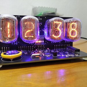 May include: A digital nixie tube clock featuring four glass tubes displaying the orange glowing numbers 1, 2, 3, and 8. The clock is built on a black circuit board with purple LED backlighting, a yellow coin cell battery, and various electronic components.
