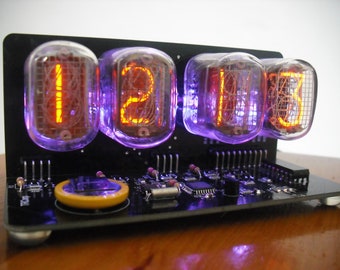 May include: A black digital clock with four glowing orange digits displayed in vacuum tubes. The tubes are encased in clear glass and emit a purple glow. The clock sits on a black circuit board base.
