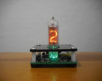 May include: A retro-style digital display featuring a glowing orange number '2' inside a glass tube. The tube sits atop a black platform with silver corner accents, mounted on a green circuit board. The display emits a soft green light.