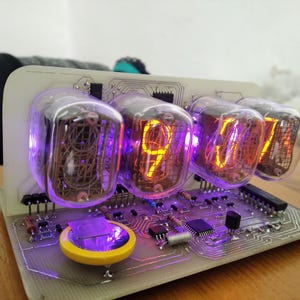 May include: A digital clock with four glowing Nixie tubes displaying the time. The tubes are encased in clear glass and emit an orange light. The clock's circuit board has various electronic components.
