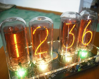 May include: A digital clock with four glowing orange Nixie tubes displaying the time 12:36. The tubes are encased in glass and mounted on a circuit board with green indicator lights. The clock is on a wooden surface.