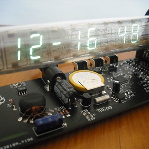 May include: A digital clock displaying 12:16:48 in bright green numerals. The clock is housed in a clear glass tube and mounted on a black circuit board with various electronic components. The board has a paw print logo and the text "TRCorp".