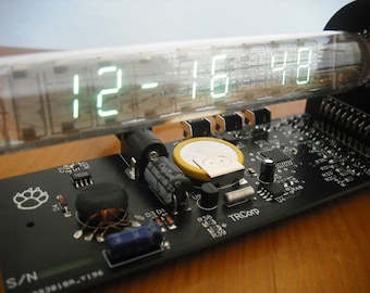 May include: A digital clock displaying 12:16:48 in bright green numerals. The clock is housed in a clear glass tube and mounted on a black circuit board with various electronic components. The board has a paw print logo and the text "TRCorp".