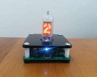 May include: A Nixie tube display showing the number 2 in orange, mounted on a black square base with silver screws. The base sits on a green circuit board with white supports. A blue light emanates from the base.