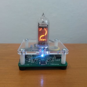 May include: A digital clock with a glowing orange number "2" displayed in a glass tube. The clock is mounted on a green circuit board base with a clear acrylic cover. The base has the text "TRCorp".