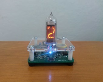 May include: A digital clock with a glowing orange number "2" displayed in a glass tube. The clock is mounted on a green circuit board base with a clear acrylic cover. The base has the text "TRCorp".