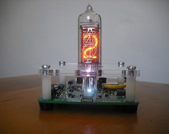 May include: A retro-style digital clock with a glowing orange display. The clock features a clear glass tube with an illuminated number, mounted on a circuit board base with a clear acrylic frame. The clock is a unique piece of technology.