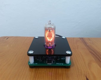 May include: A glowing orange Nixie tube display, encased in a black acrylic base with silver screws. The display sits on a green circuit board, with the text "TRCorp" visible. The tube emits a warm, inviting light.