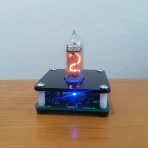 May include: A Nixie tube display showing the number 2, encased in a glass tube. The tube glows orange, mounted on a black and green base with a blue LED light. The base is supported by white cylindrical posts.