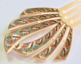 May include: A cream-colored hair comb featuring a fan-shaped head with five prongs. The surface is decorated with intricate gold-toned scrollwork, vibrant red, blue, and green enamel accents, and small embedded dark red rhinestones in a pattern.
