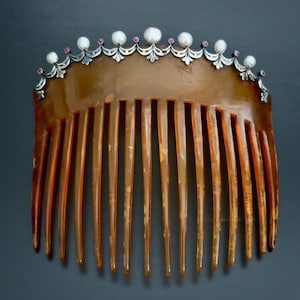 May include: A vintage hair comb with a decorative top. The comb is made of a translucent brown material with long, vertical teeth. The top is adorned with white pearls and small pink gemstones set in a silver-colored metal.