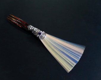 Puede incluir: Un peine de pelo de plata con una piedra preciosa azul y un diseño decorativo. El peine tiene un mango largo y delgado con un cepillo de fibras blancas, azules y rosas.