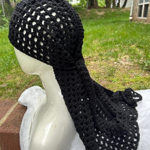 May include: Black crochet head covering with a long, flowing tail. The head covering is made of a net-like weave, allowing glimpses of the mannequin head beneath. The tail is knotted at the base of the head covering. The head covering is displayed on a white mannequin head.