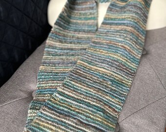 May include: A knitted scarf in shades of blue, green, and tan. The scarf is displayed on a mannequin and has a striped pattern. The scarf is made of a soft, textured yarn and appears to be handmade.
