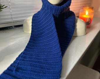 May include: A handmade, royal blue crochet scarf is draped around a white mannequin head. The scarf has a ribbed texture and a turtleneck design. A lit candle is visible in the background, adding warmth to the scene.