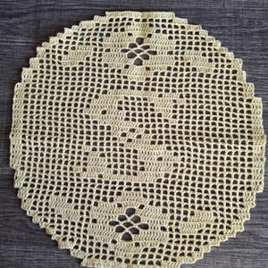 May include: A round, cream-colored crocheted doily with a delicate openwork design. The doily features a repeating pattern of small squares and floral motifs, creating a textured surface. The edges are scalloped, adding a decorative touch to this handmade item.