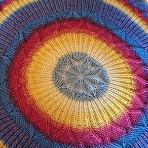 May include: Circular, hand-knitted blanket in a vibrant rainbow of colors. The design features concentric rings of blue, red, yellow, and purple, with a central starburst pattern. The blanket is made of soft yarn and has a textured, tactile appearance.