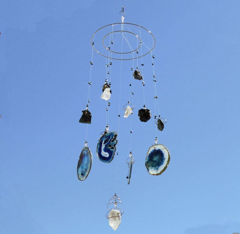 May include: A hanging mobile with a circular metal frame, featuring various natural stones and crystals suspended by strings. The stones include dark, light, and blue-toned slices, and clear crystals. The mobile hangs against a clear blue sky.