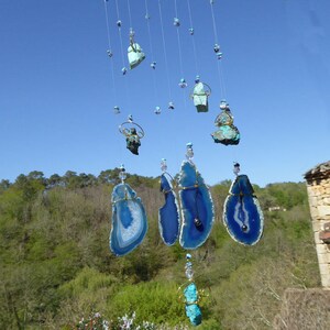 May include: A hanging wind chime featuring various blue and turquoise stones and agate slices. The agate slices are edged with gold wire and strung with clear beads. The background shows a clear blue sky and green foliage.