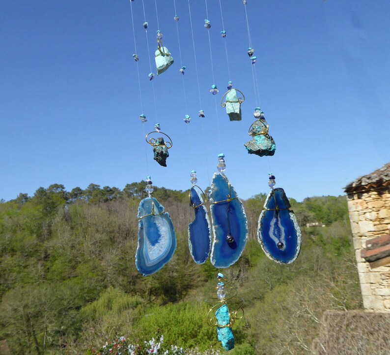 May include: A hanging wind chime featuring various blue and turquoise stones and agate slices. The agate slices are edged with gold wire and strung with clear beads. The background shows a clear blue sky and green foliage.